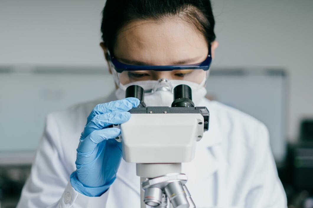A researcher wearing protective gear analyzes a sample through a microscope in a laboratory.