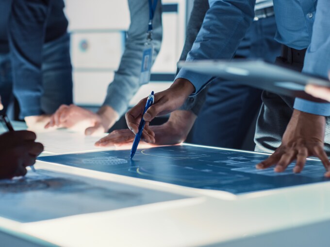 Hands and tools of an engineering team discussing over a blueprint spread out on a brightly lit table.