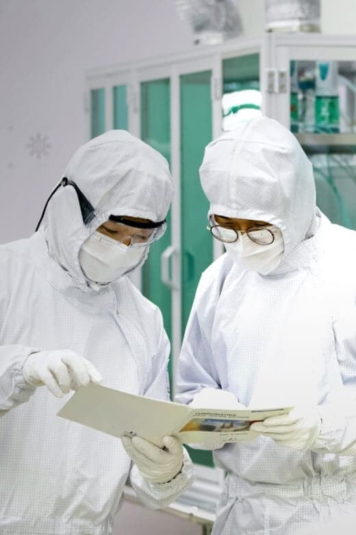 防塵服を着用した2人の研究員がクリーンルームで文書を検討している様子です。